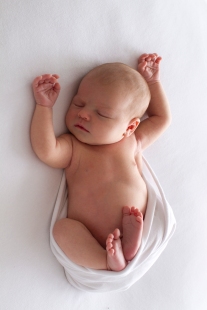Newborn photography, baby photographed in studio, on white fabric, wrapped in white wrap. Baby look relaxed with arms above head, feet curled up.